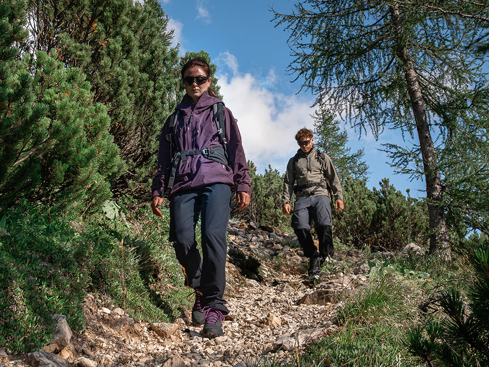 Two hikers descend a rocky forest trail under a blue sky wearing SCARPA Ribelle Cross 2 Mid GTX light hiking boots.