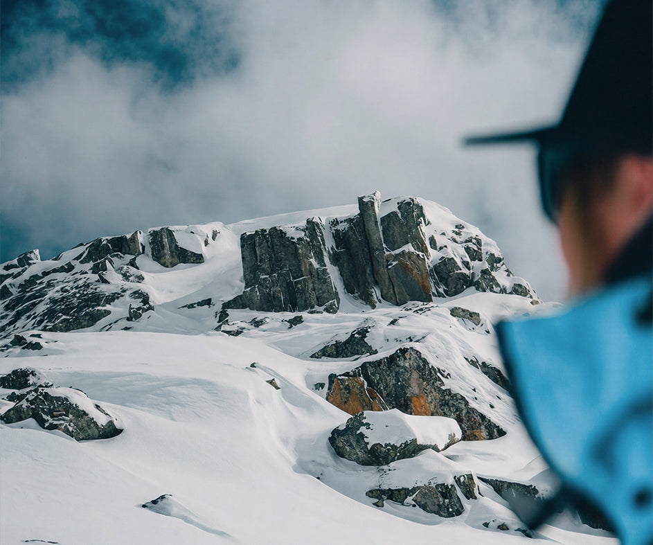 A snowy mountain peak with dark rocky cliffs under a cloudy blue sky, with the blurred side of a skier wearing SCARPA's Gea ski mountaineering boots.